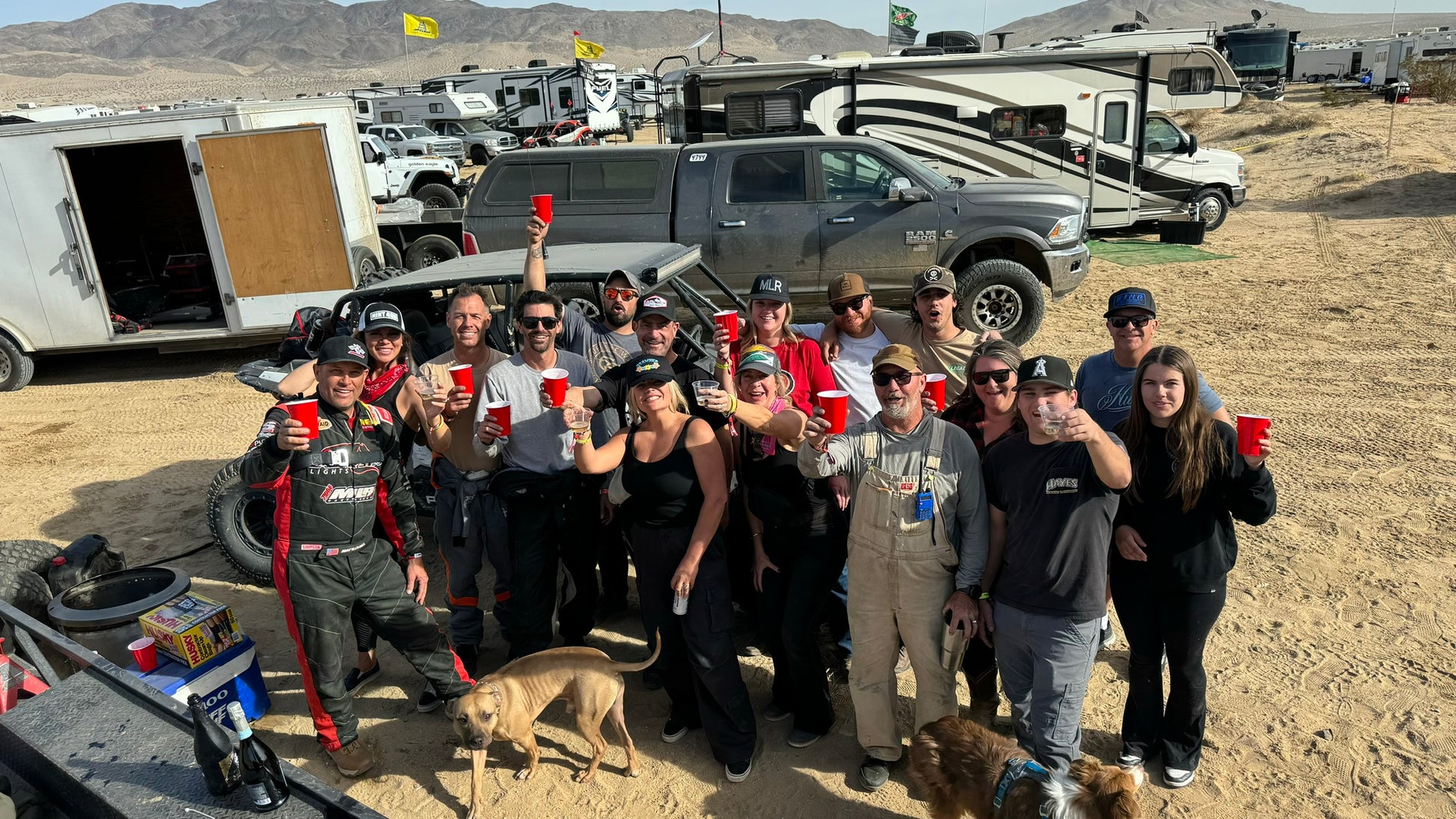 Group of people and dogs posing for a photo outdoors with vehicles and mountains in the background