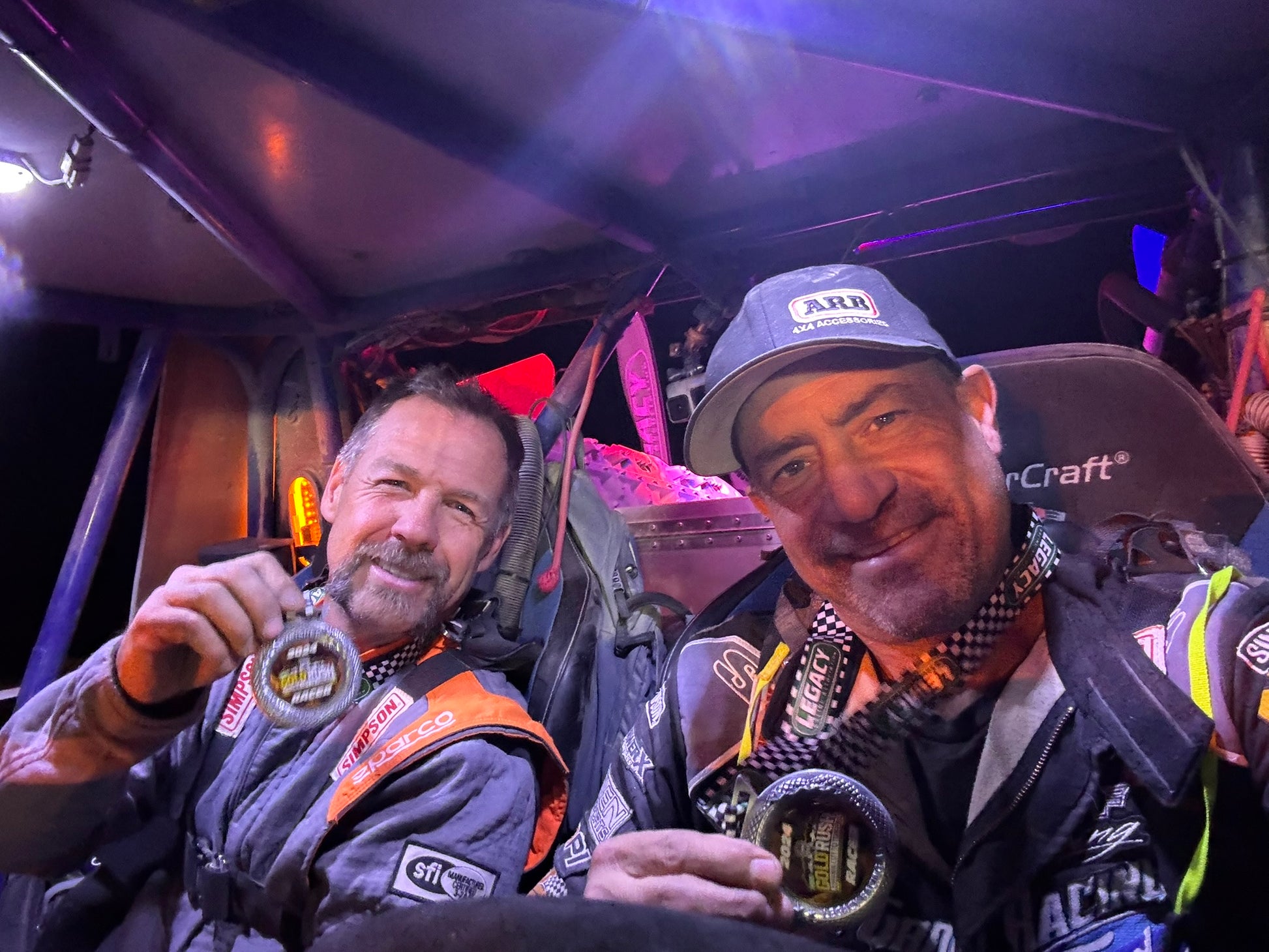 Two men inside a vehicle, holding awards, with a dark interior background.